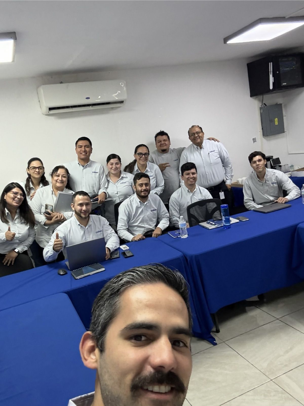 Rogelio en primer plano con el equipo de Zubex, unas catorce personas, en su sala de juntas, con laptops sobre mesas con manteles azules